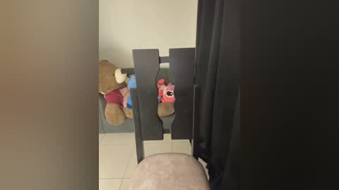 A video of a beige room with a black chair and two stuffed animals: a brown bear and a pink bunny. The background features light gray walls and a tiled floor.