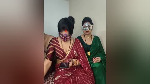 Video of two women in traditional Indian attire, one in red with gold jewelry, the other in green, seated on a couch with blurred background.