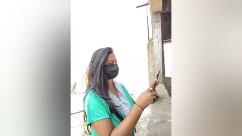 Video of a South Asian woman with long black hair, wearing a blue mask and green shirt, sitting on a concrete step, holding a smartphone, looking outdoors.