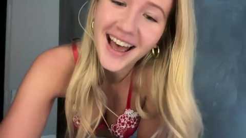A video of a smiling, blonde Caucasian woman with fair skin, wearing a red and white floral-patterned bra, and large hoop earrings, leaning forward.