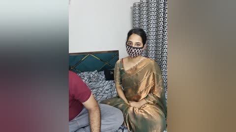A video of a woman in a gold sari and face mask, seated on a bed with patterned bedding, next to a man in a red shirt.