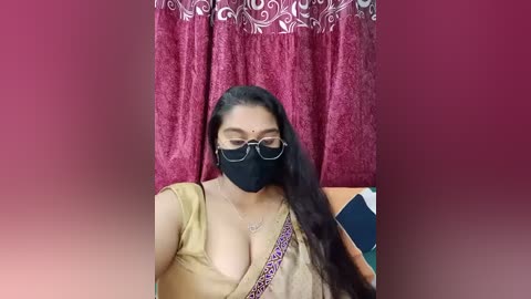 Media: A video of a South Asian woman with long black hair, wearing glasses, a black mask, and a beige blouse with a leopard print shoulder strap, standing in front of a red curtain with white floral patterns.