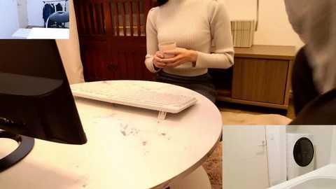 Video of a woman in a cream sweater, standing at a white marble-topped table with a computer and wooden furniture in a cozy, well-lit room.