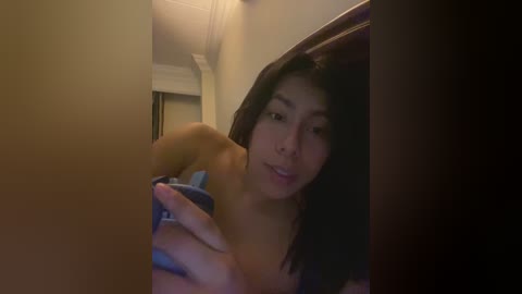 Video of a young Asian woman with medium-length black hair, wearing a blue bra, taking a selfie in a dimly lit room with beige walls and wooden furniture.