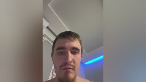 A video of a shirtless man with short brown hair, light skin, and a trimmed beard, standing in a dimly lit room with white walls, a blue LED strip, and a partially visible ceiling fan.