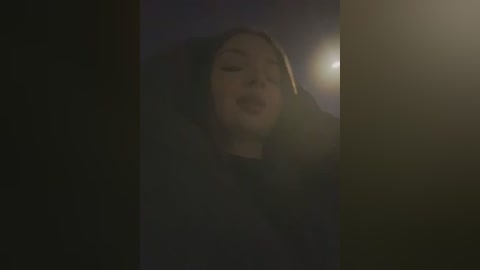 A dimly lit video of a woman with long blonde hair, wearing a black hooded cloak, her face partially obscured by shadows, standing in a dark, shadowy room.