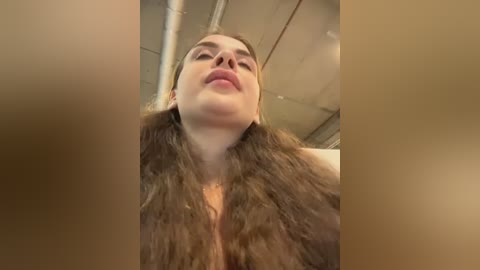 A close-up video of a woman with long, wavy brown hair and fair skin, wearing a fur coat, standing in a dimly lit room with industrial ceilings.