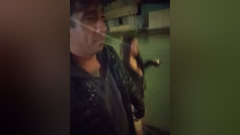 Video of a man with a dark, leather jacket and glasses, partially blurred, standing in a dimly lit, green-tiled bathroom with a toilet in the background.