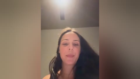 Media: A video of a woman with long, straight black hair, light skin, and closed eyes, standing in a dimly lit room with a ceiling light shining directly above her head.