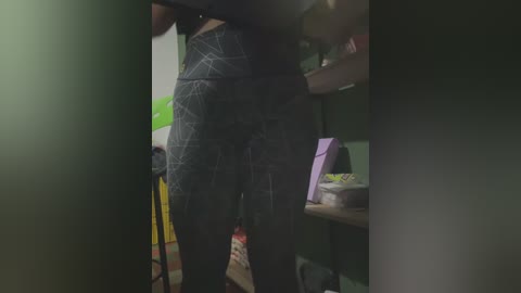A video of a woman in black patterned leggings, standing in a dimly lit room, with a green wall and a purple bag on a wooden table in the background.