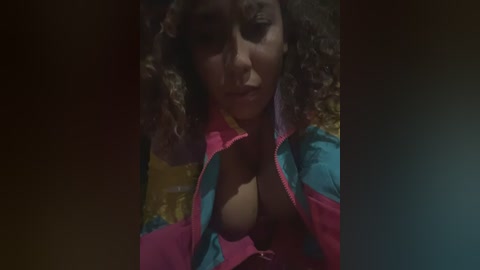 Video of a dark-skinned woman with curly hair, wearing a bright pink and blue jacket, exposing cleavage, in a dimly lit, possibly outdoor setting.
