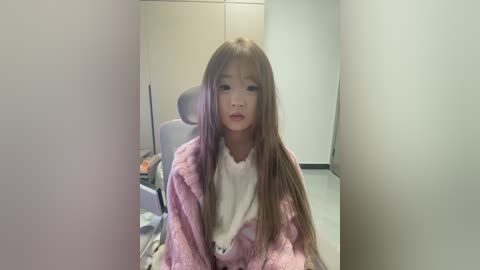 Video of a young Asian girl with long brown hair, wearing a fluffy pink bathrobe over a white shirt, sitting in a modern, minimalistic bathroom with beige walls and a gray chair.