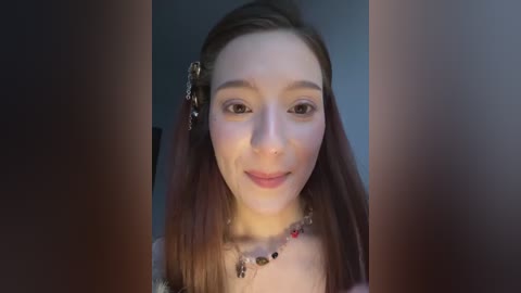 A video of a young woman with fair skin, light brown hair, and a slight smile, wearing a beaded necklace, against a dark background.