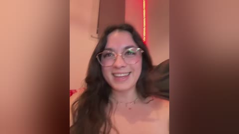 A video of a smiling young woman with long brown hair, wearing glasses, and a strapless top, standing in a dimly lit room with red lighting.