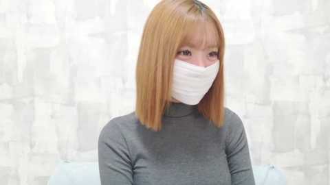 Media: Video of a young Asian woman with straight, shoulder-length, light brown hair and fair skin, wearing a grey turtleneck sweater and a white face mask. She stands against a white and grey patterned wall.
