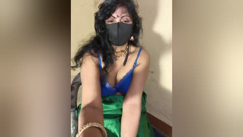 Video of a South Asian woman with medium skin tone, long black hair, wearing a blue bra, black face mask, gold necklace, and green cloth, seated indoors.