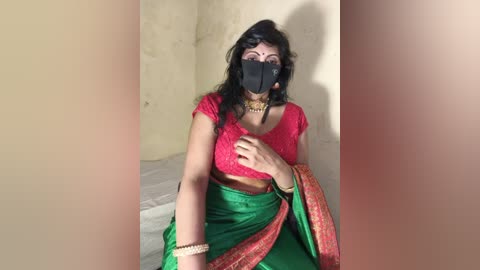 Video of a woman in traditional Indian attire, a red lace blouse, green and gold sari, and black mask, seated on a white bed against a plain beige wall, wearing a gold necklace.