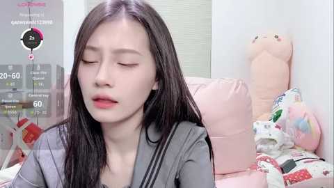 Video of an East Asian woman with long black hair, closed eyes, and pale skin, sitting on a pink couch in a room with stuffed animals.