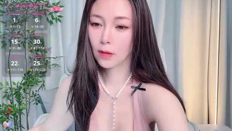 A video of a young East Asian woman with long, straight black hair and fair skin, wearing a low-cut pink top with a black cross necklace, in a softly lit room with floral decor and a digital clock overlay.