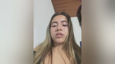 Video of a young woman with long, straight brown hair, light skin, and a neutral facial expression, wearing a black top. Background features a wooden ceiling and plain white walls.