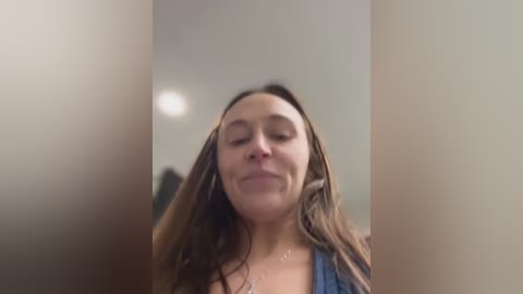 A video of a young woman with long, wet hair, closed eyes, and a slight smile, wearing a blue top. She stands in a dimly lit bathroom with beige walls and a blurry background.