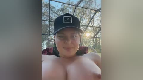 Video of a fair-skinned, topless woman with large breasts, wearing a black baseball cap, sitting on a red-patterned chair inside a glass-walled room with a forest background at sunset.