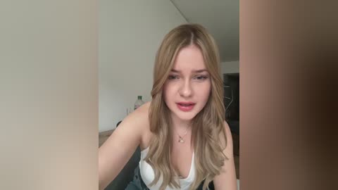 Video of a young Caucasian woman with long, wavy blonde hair, wearing a white tank top, and smiling softly, set indoors with a neutral background.