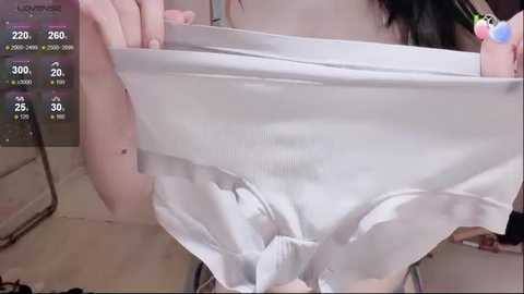 A close-up video of a person's hand holding up white underwear, revealing a smooth, pale thigh. The background shows a bathroom with a mirror and a scale displaying 220 pounds.