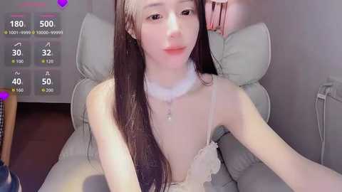 A young Asian woman with long, straight black hair and fair skin poses in a white, frilly lingerie top, sitting on a light-gray cushioned chair. The background features a purple heart overlay and a digital display with metrics.