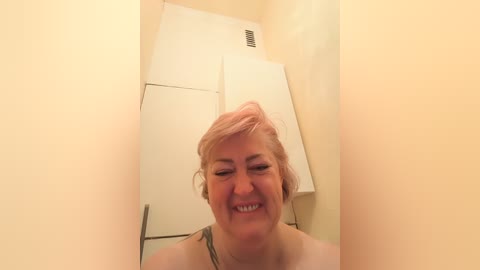 Video of a smiling, middle-aged woman with short, pinkish-blonde hair in a small, beige-walled room with a white, metal door and a small window. She wears a light-colored top.