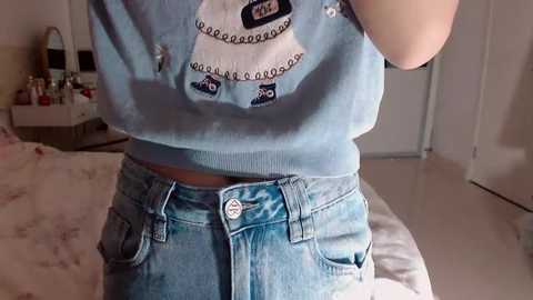 Video of a young woman wearing a light blue sweater with a cartoon panda design and high-waisted, light blue denim shorts. The background features a bed with floral bedding, a dresser, and a closed door.