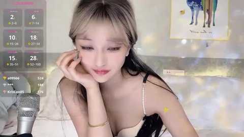 Video of a young Asian woman with long, straight, blonde hair, wearing a black and white striped top, lying on a bed, looking contemplative. Background includes a poster and a mic.