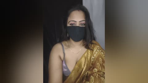 Video of a South Asian woman with medium skin tone, straight black hair, wearing a black face mask, mustard yellow sari with floral pattern, and a pink bra, in a dimly lit room with a blurred background.