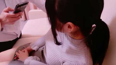A video shows a young woman in a light gray sweater, lying on a white bed, looking at her phone, while a doctor in a white coat stands by.
