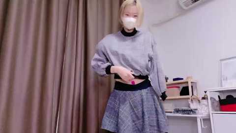 Video of a woman with short blonde hair, wearing a gray checkered dress, black choker, and white mask, in a sterile room with beige curtains, shelves, and a white wall.