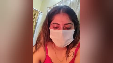 Video of a young woman with long brown hair, wearing a white surgical mask and a red lace bra, standing in a bathroom with string lights, yellow curtain, and a towel rack.