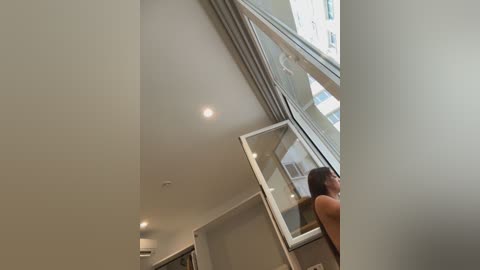 Video of a modern, minimalist room with a large glass wall, showcasing a person with dark hair and bare shoulders standing in front of it.