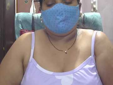 Video of a South Asian woman with light brown skin, wearing a blue surgical mask, light purple spaghetti strap top, and gold necklace, seated on a teal chair in a home setting.