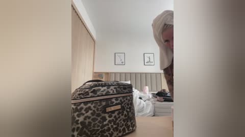Video of a woman in a white towel, standing in a tidy, beige-tiled bathroom with a leopard-patterned cosmetic bag, framed art, and a neatly made bed in the background.