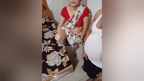 Video of an elderly Indian woman in a floral sari, red blouse, and white dupatta, sitting on a bed with a floral bedspread, in a modest, tiled room.