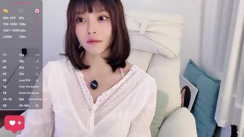 Video of a young Asian woman with straight, shoulder-length brown hair, wearing a white lace blouse, sitting on a white couch in a minimalist room.