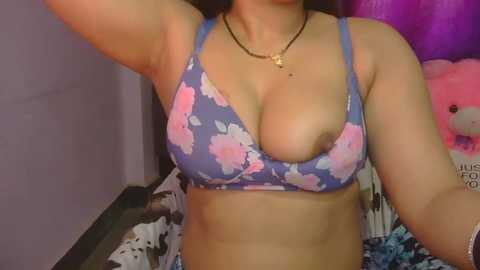 Video of a light-skinned woman with medium-sized breasts, wearing a blue floral bra and a black chain necklace, in a purple room with stuffed toys.