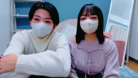 Video of two Asian women in casual, pastel-colored sweaters with face masks, seated indoors. Background features a light blue wall, white shelving unit, and a pink armchair.