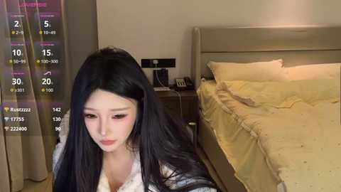 A video of an Asian woman with long black hair, fair skin, and pink lipstick, sitting on a bed in a modern bedroom with yellow sheets and beige walls.