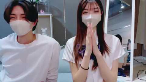 Video of two young Asian women with pale skin, wearing white masks and school uniforms, hands clasped in prayer. Background shows a modern, minimalist room with a shelf and a desk.