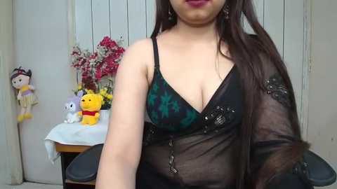 A video of a woman with long, dark hair, wearing a sheer black blouse with a green tie-dye bralette underneath, seated indoors. Background includes a stuffed toy, a flower arrangement, and a wooden table.