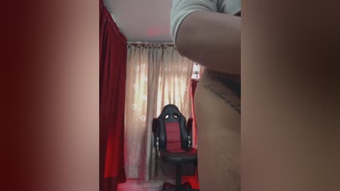 Video of a narrow, dimly lit room with red curtains on one side, beige curtains in the back, and a black gaming chair centered, with a person's arm visible in the foreground.