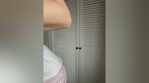 Video of a partially obscured person in a white shirt and pink checkered skirt, standing in front of white louvered closet doors with black knobs. The scene is dimly lit.