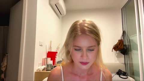 Video of a blonde woman with fair skin, wearing a white tank top, standing in a modern, minimalist room with white walls, an air conditioner, and a glass door.