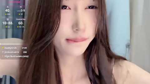 Media: A close-up video of an Asian woman with long brown hair, fair skin, and a slight smile, wearing a sleeveless top. The background is blurred, featuring a window and a light-colored wall.